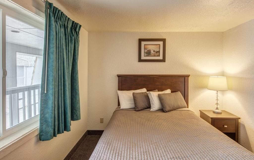 A hotel room with a bed, a nightstand with a lamp, a window with curtains, and a picture on the wall.