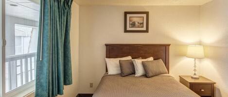 A hotel room with a bed, a nightstand with a lamp, a window with curtains, and a picture on the wall.