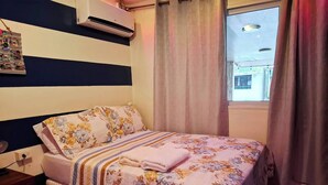 Een slaapkamer met een bed, een nachtkastje, een wandairconditioner, een raam met gordijnen en uitzicht op buiten.