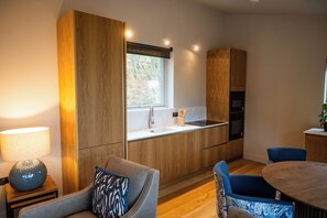 Une cuisine moderne avec des meubles en bois, un évier encastré et une fenêtre. On y trouve une table de salle à manger ronde, des chaises bleues et une lampe sur une petite table d’appoint.