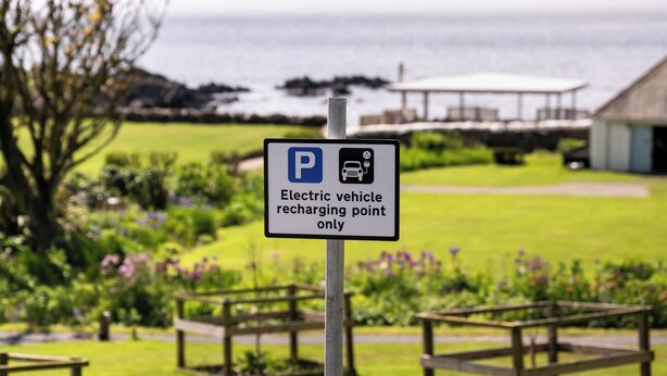 Señal de punto de carga para vehículos eléctricos en un parque con mesas de picnic y vistas al mar.