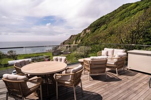 Una terraza con mobiliario de mimbre, una mesa redonda y vistas al mar y al verdor.