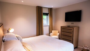 Ein Hotelzimmer mit einem Bett, einem an der Wand montierten Fernseher, einem Schminktisch, einem Sessel und einem Fenster mit Vorhängen.