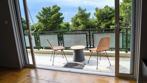 A balcony with a table and two chairs, a view of trees and a sailboat, and a wooden floor inside.