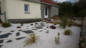 Una casa amb un camí de pedra, un jardí petit amb diverses plantes i una terrassa de fusta.