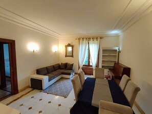A living room with a sectional sofa, a dining table with chairs, a small side table, a bookshelf, and a window with curtains. - FRGK OXFORD palace park (Cannes)
