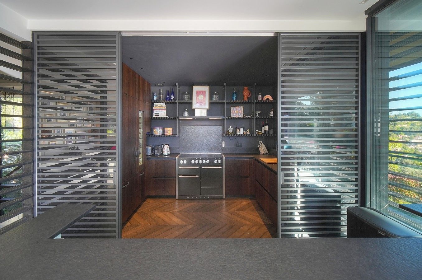 Cozinha moderna com piso de madeira em padrão herringbone, fogão de aço inoxidável e vista através de amplas janelas.