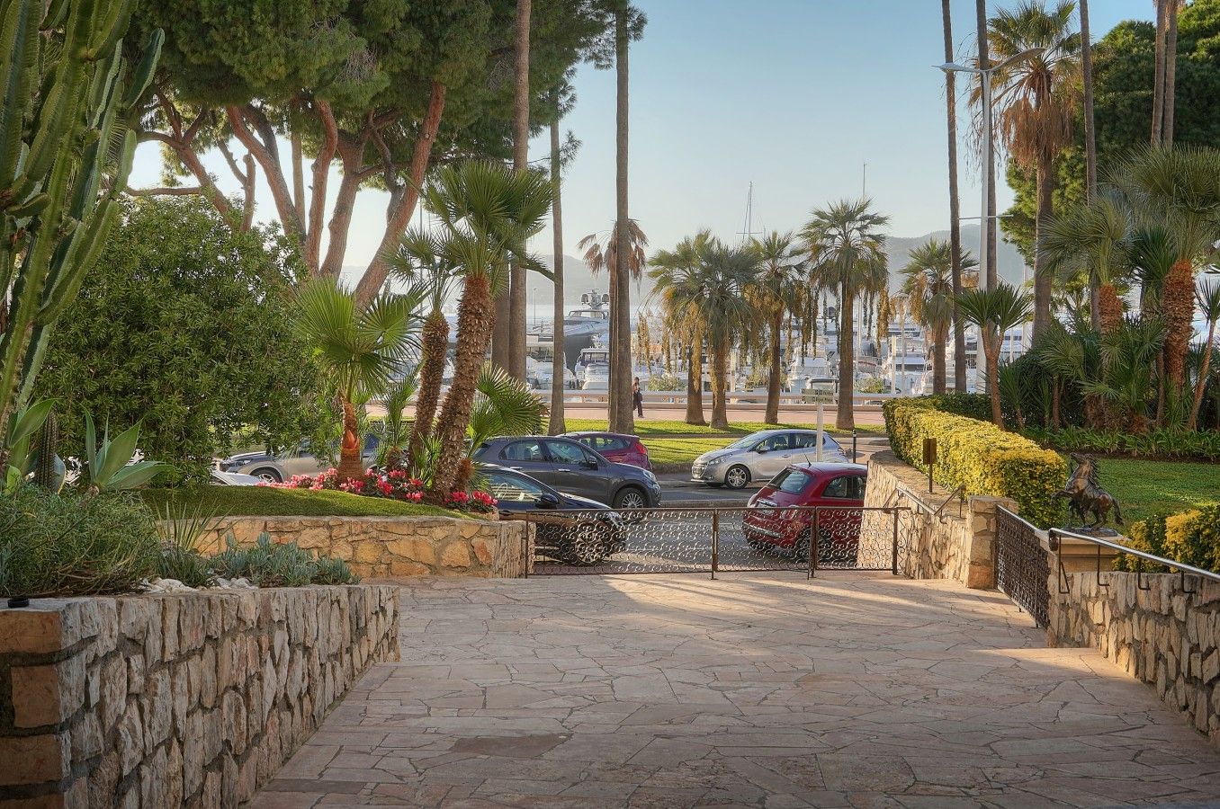 A paved pathway leading to a gated entrance with parked cars and palm trees.