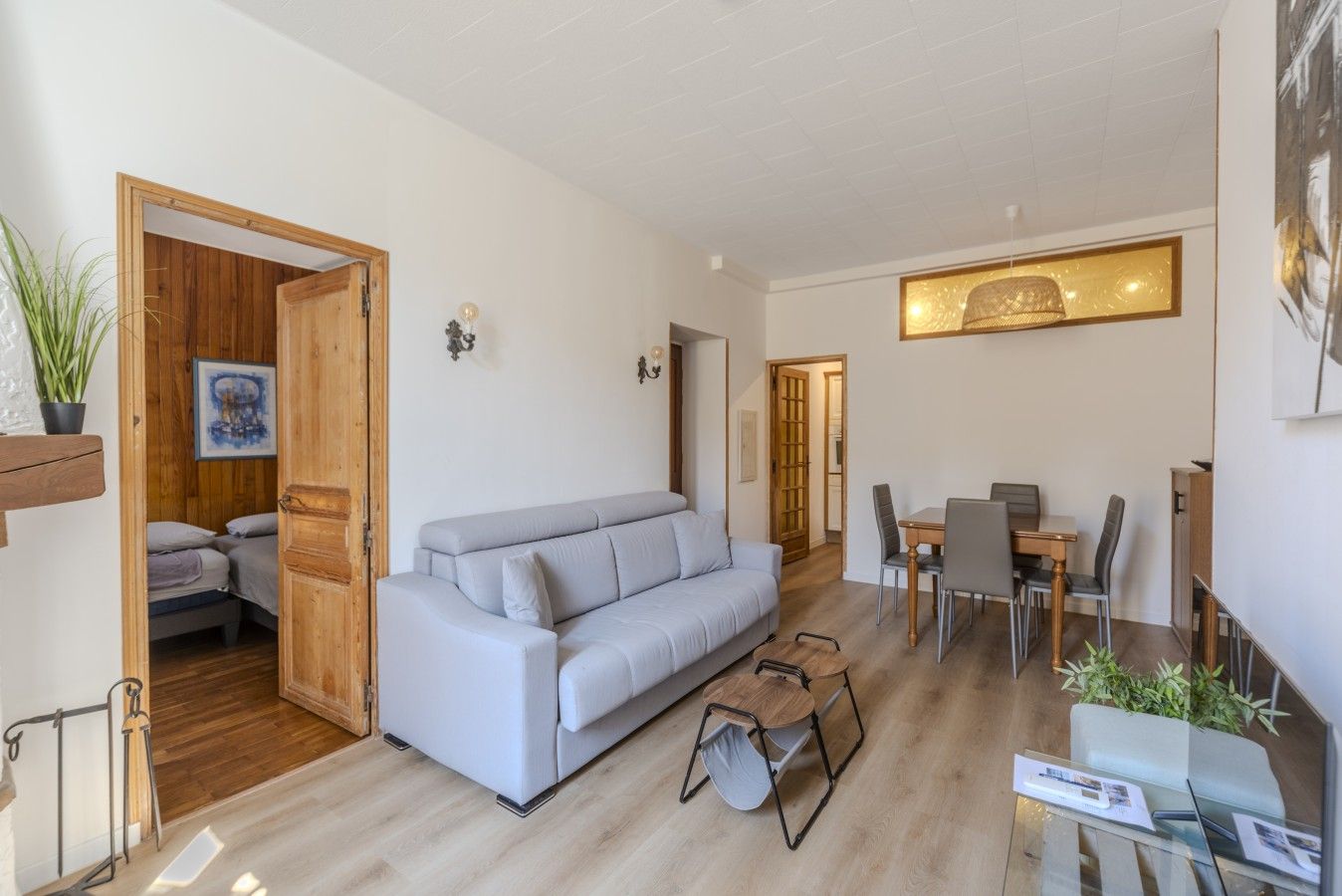 A modern living room with a sofa, dining table, and chairs. There is a wooden door leading to a bedroom.