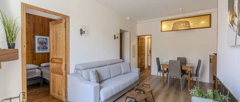 A modern living room with a sofa, dining table, and chairs. There is a wooden door leading to a bedroom.