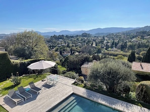 Ein Poolbereich mit Liegestühlen und Sonnenschirm, umgeben von gepflegten Gärten und Blick auf ein Wohngebiet mit Hügeln im Hintergrund.