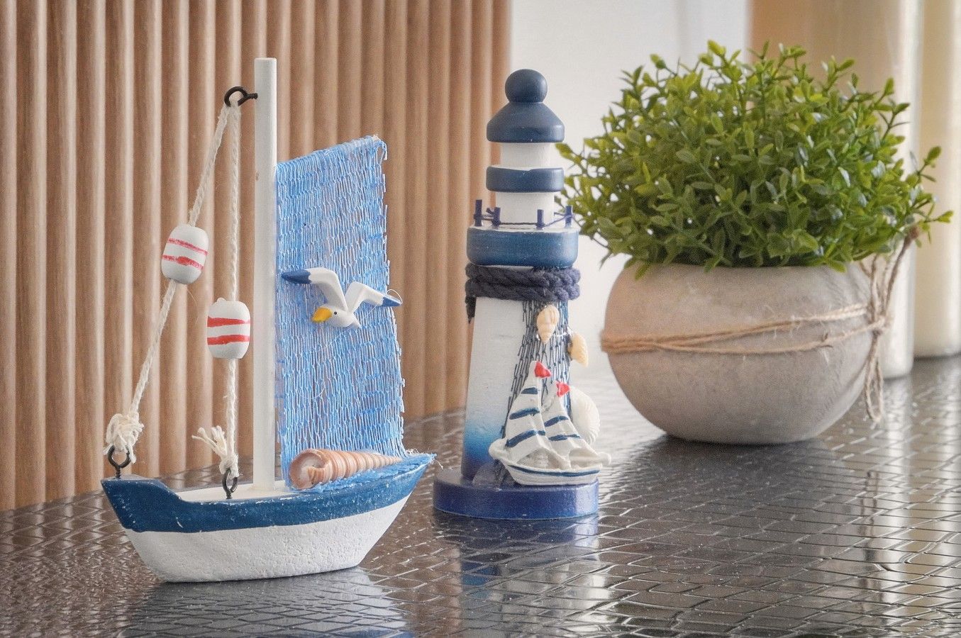 Un petit voilier, un phare et une plante en pot, le tout posé sur une surface carrelée.