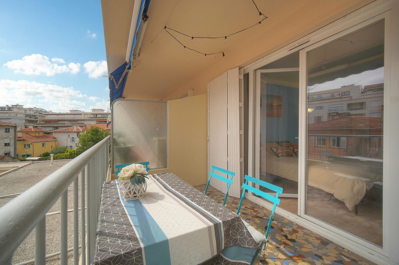 Een balkon met een tafel en stoelen, een bloevas en een uitzicht op gebouwen door een glazen deur.