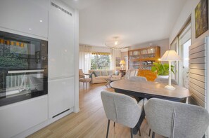 A modern kitchen with built-in appliances, a dining area with a table and chairs, and a living room with a sofa and a bookshelf. - 10046194 FRGK "Prince of Wales Garden Terrace" (Cannes)