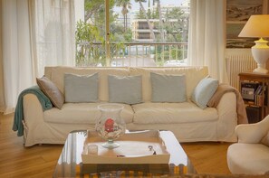 A living room with a beige sofa, a glass coffee table, and a view of a balcony with plants and buildings. - 10046194 FRGK `Prince of Wales Garden Terrace` (Cannes)
