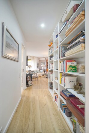 Ein Flur mit Bücherregal, gefüllt mit Büchern und Dekoration, der in einen Wohnbereich mit Sofa und Lampe führt.