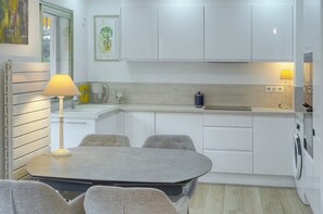 A compact kitchen with white cabinets, a dining table with chairs, a lamp, and a washing machine. - 10046194 FRGK `Prince of Wales Garden Terrace` (Cannes)