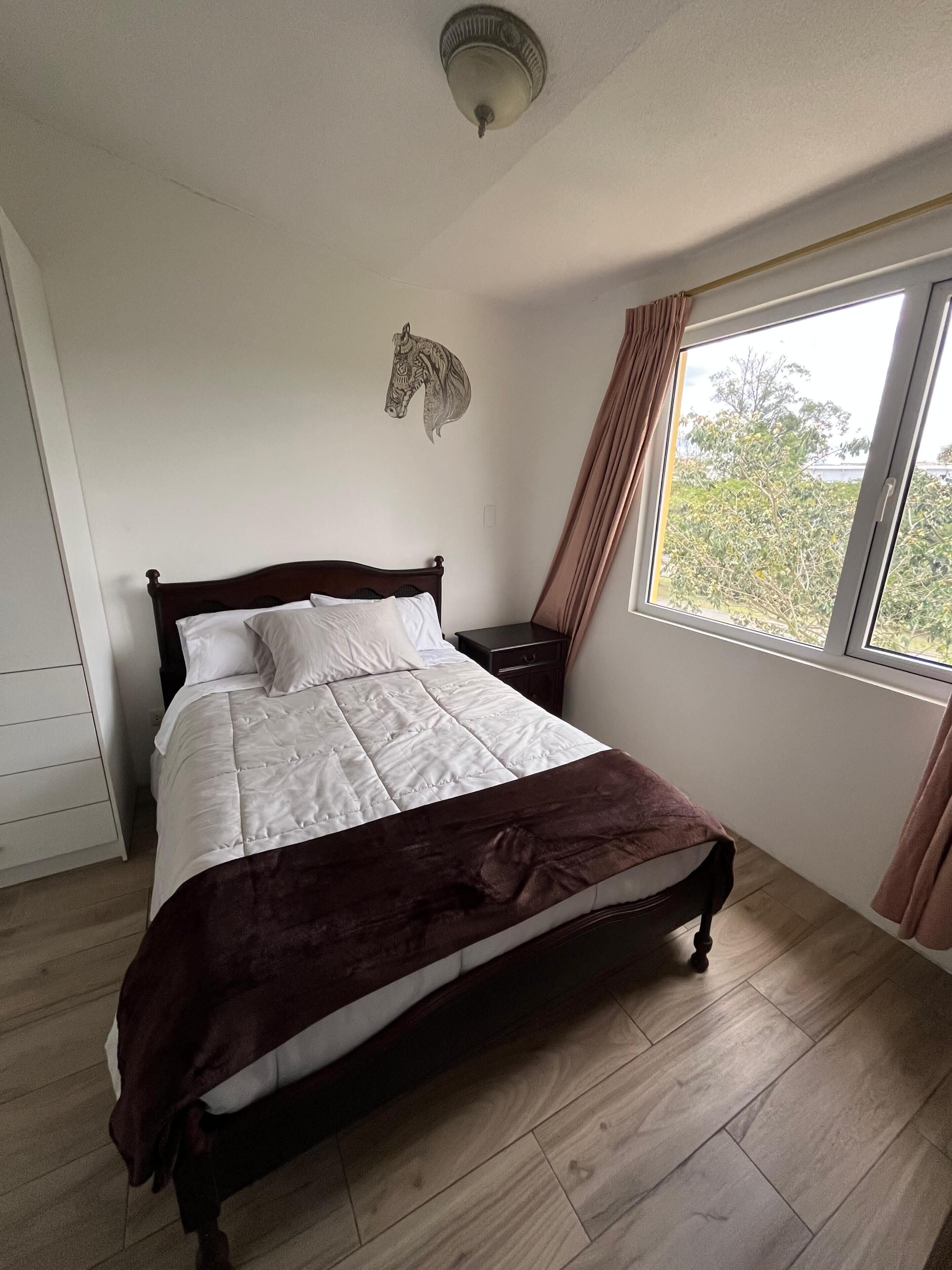 A bedroom with a bed, a nightstand, a ceiling fan, a window with curtains, and a wall-mounted horse decal.