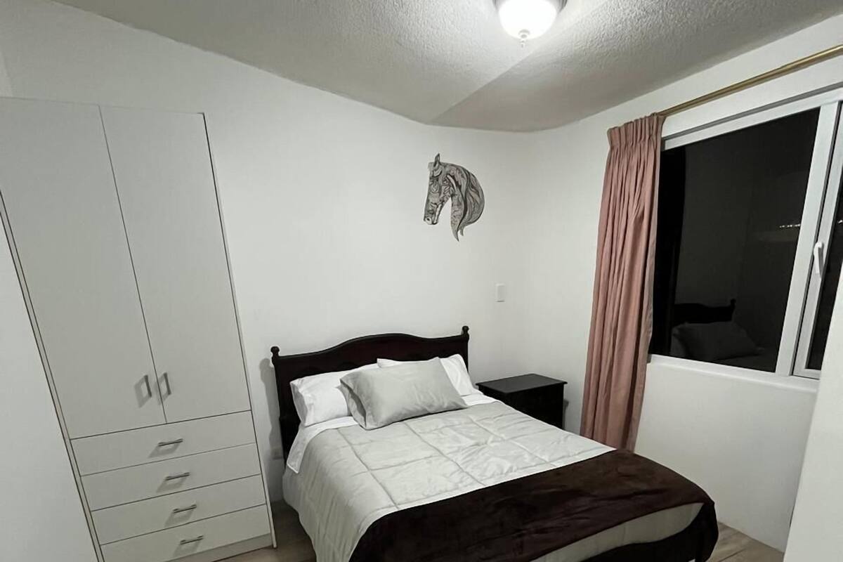 Ein Schlafzimmer mit Bett, Kleiderschrank, Nachttisch, einem Fenster mit Vorhängen und einer Deckenleuchte.