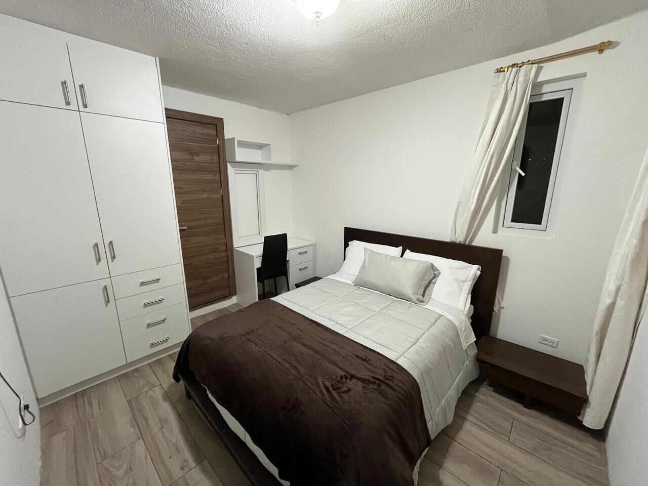 Un dormitorio con cama, armario, escritorio, silla y ventana con cortinas.