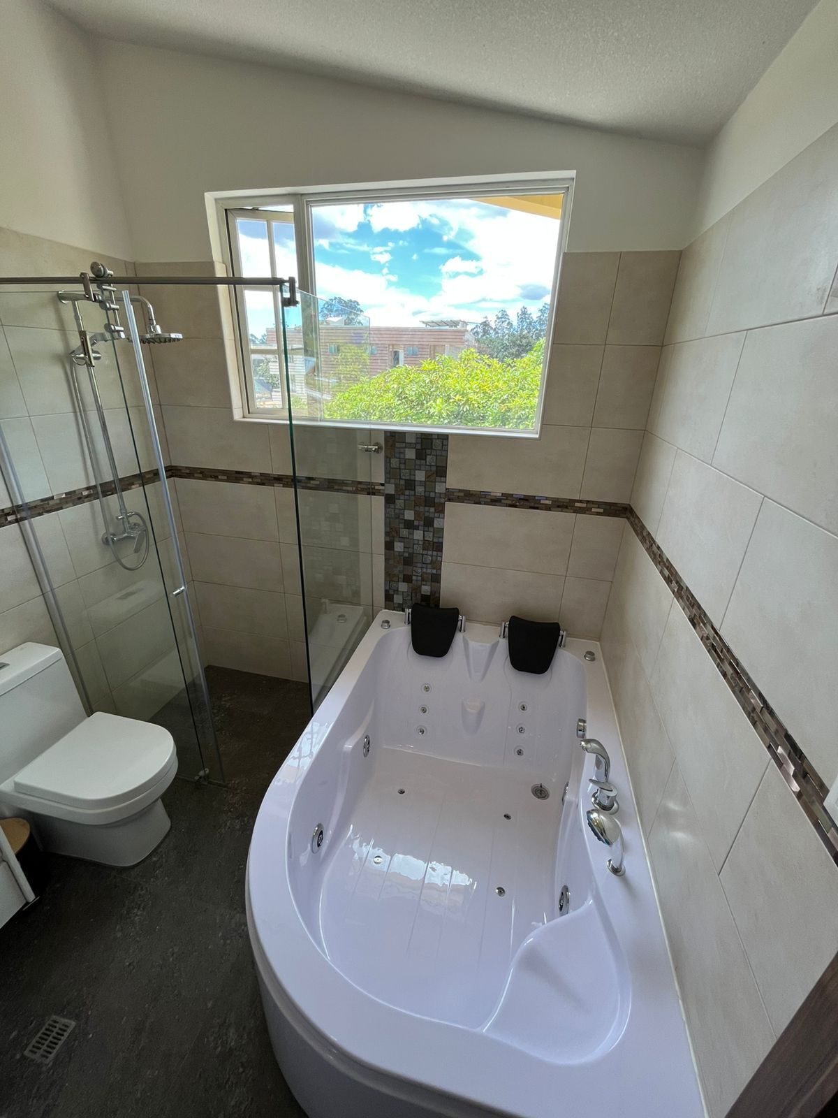 A bathroom with a glass shower enclosure, a white toilet, a bathtub with two black seats, and a window providing a view of greenery.