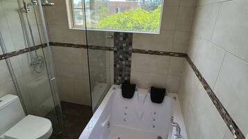A bathroom with a glass shower enclosure, a white toilet, a bathtub with two black seats, and a window providing a view of greenery.