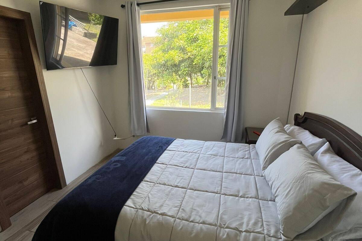 Ein Schlafzimmer mit einem Bett, einem an der Wand montierten Fernseher, einem Fenster mit Vorhängen und einer Tür.