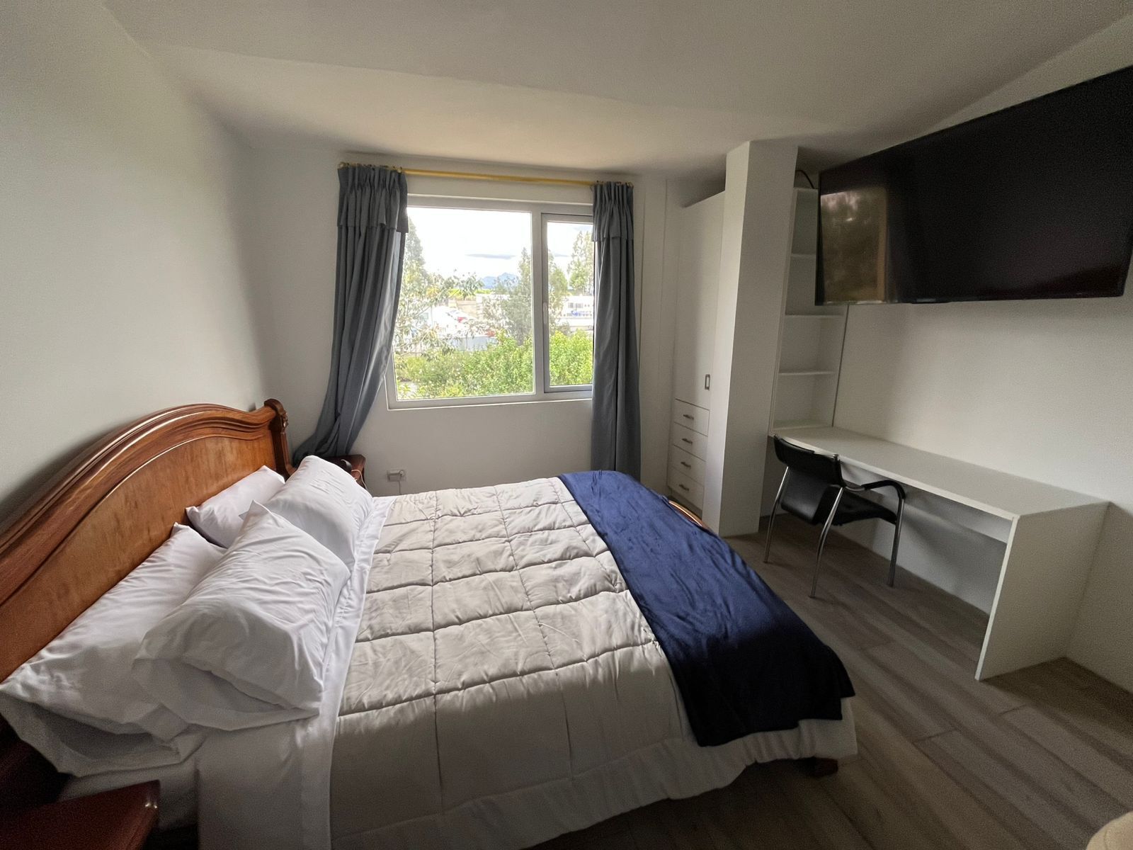 A bedroom with a bed, a desk, a chair, a TV, and a window with curtains.
