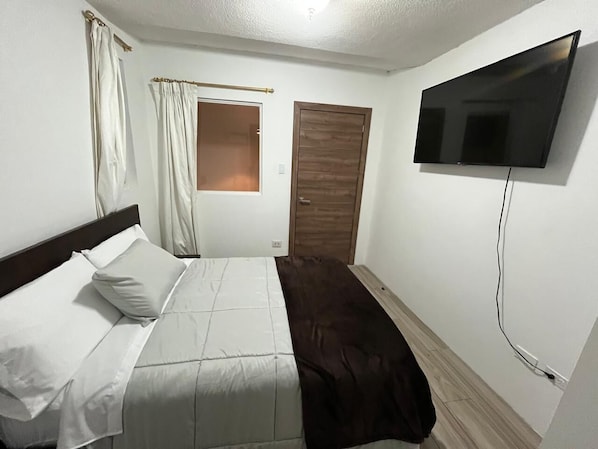 A bedroom with a bed, a window with curtains, a door, and a TV mounted on the wall. - Hotel Arupo Residence Aeropuerto (Tababela)