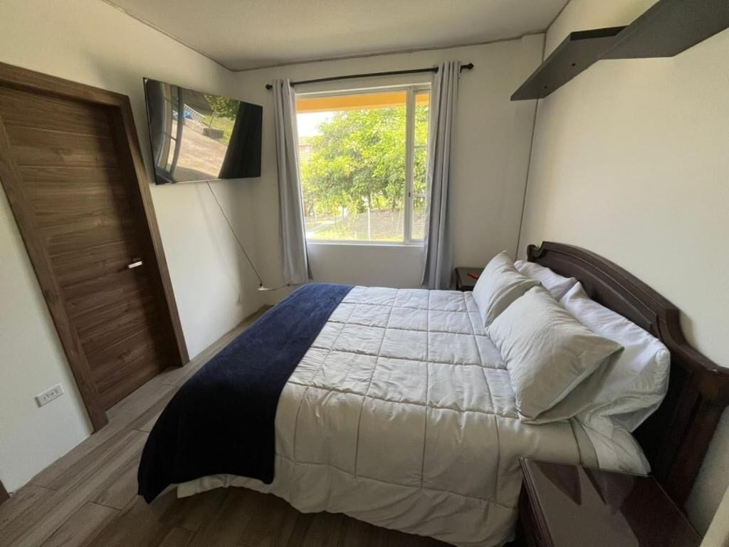 Un dormitorio con una cama, televisor montado en la pared, una ventana con cortinas y una puerta de madera.