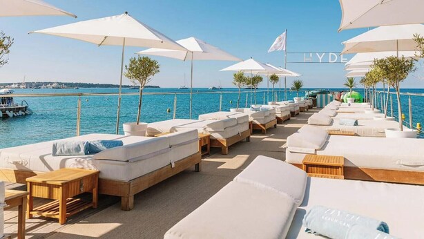 A beachside lounge area with rows of white lounge chairs and umbrellas, a wooden deck, and a view of the sea. - SURCOUF (Cannes)