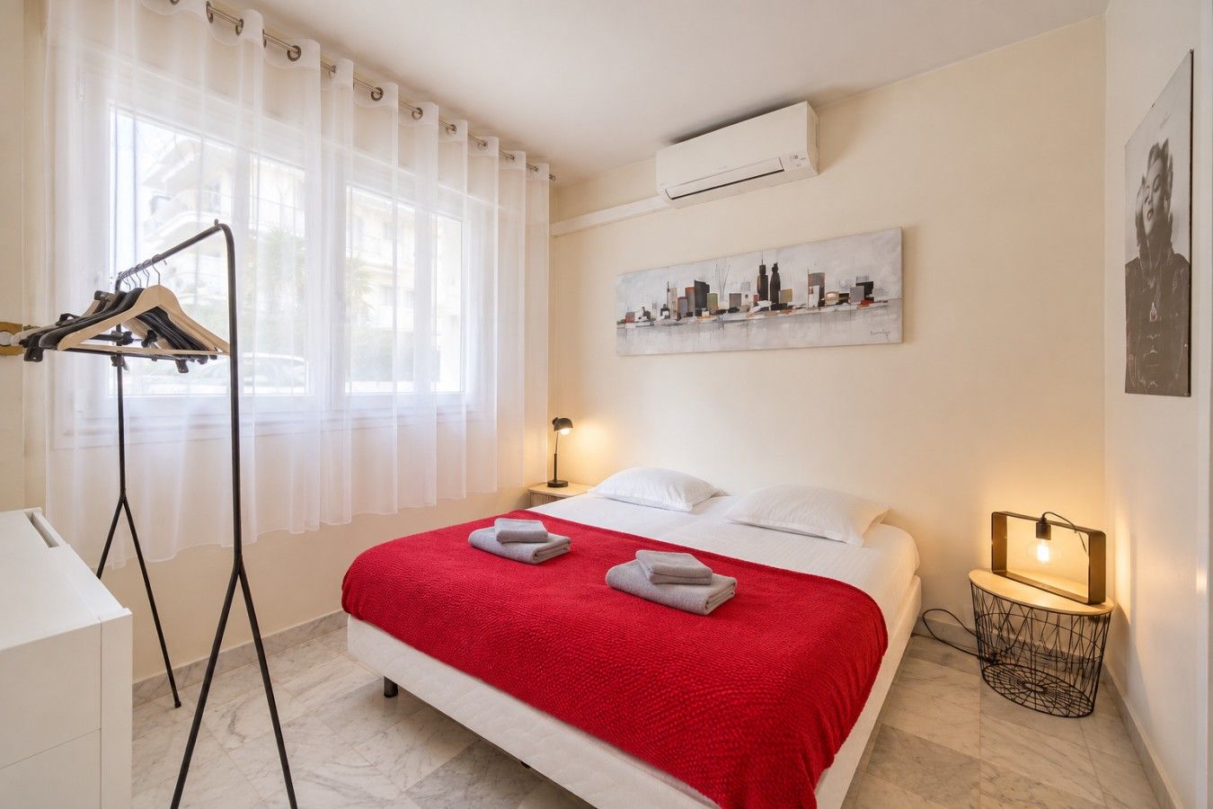 Habitación de hotel con una cama, una lámpara de noche, un aire acondicionado de pared y un cuadro con una vista urbana.