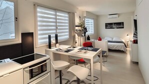 A modern, compact hotel room with a kitchenette, a dining area, a bed, and a large window with blinds. - SURCOUF (Cannes)