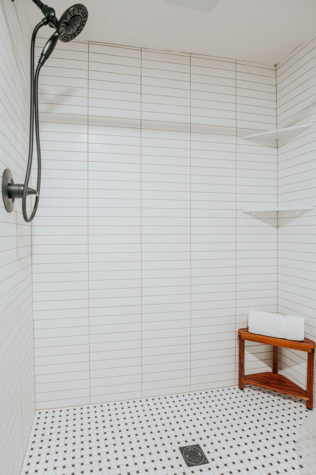 A shower with a handheld showerhead, a wooden bench with a towel, and a tiled floor with a drain.