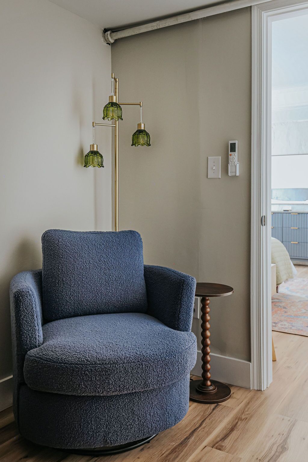 Habitación de esquina con un sillón azul, una lámpara de pie, una mesita redonda y un ventanal corredero que da a un balcón.