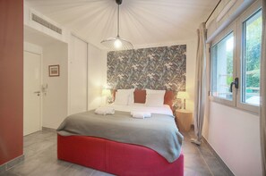 A bedroom with a bed, a red ottoman, a wall with a floral pattern, and a window with curtains. - Frgk *Diane | Elegance and Comfort in Antibes* (Antibes)