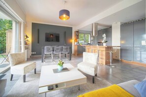 Ein modernes Wohnzimmer mit einem weißen Couchtisch, zwei Sesseln, einer Couch, einem Essbereich mit Tisch und Stühlen sowie einer Küche mit Barbereich.