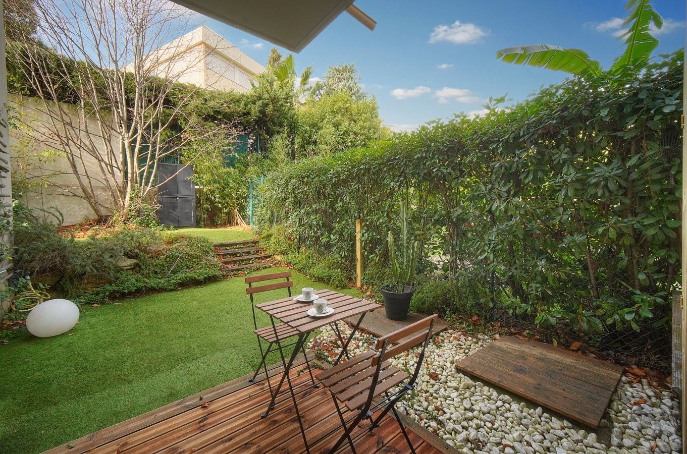 A patio with a wooden table and chairs, surrounded by a well-maintained garden with a variety of plants and a small lawn.