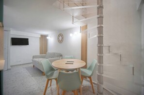 Ein modernes Wohnzimmer mit einem runden Holztisch, hellgrünen Stühlen, einer Couch und einem Flachbildfernseher, sowie einem dekorativen Spiegel.