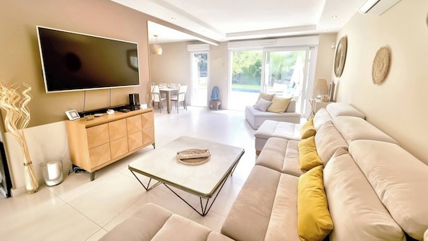 Una sala de estar moderna con un televisor de pantalla plana, un mueble de madera para el entretenimiento, un sofá color beige y una zona de comedor con una mesa y sillas.
