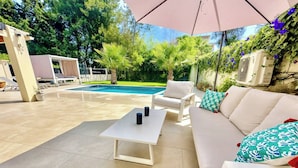 Zona junto a la piscina con un sofá blanco, dos sillas blancas, una mesa y una sombrilla.