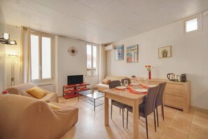 Ein Wohnzimmer mit einer Couch, einem Esstisch für zwei Personen, einem Fernseher und einer kleinen Küchenzeile mit einer Kaffeemaschine.