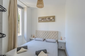 Quarto com uma cama grande, janela com cortinas, mesa de cabeceira com abajur e uma obra de arte na parede.