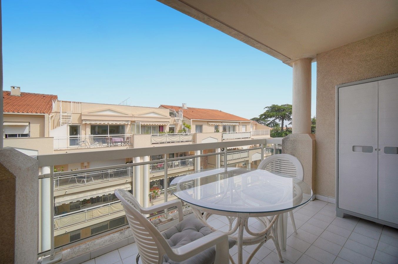 A balcony with a glass table and two chairs, overlooking a residential area with houses and a clear sky.