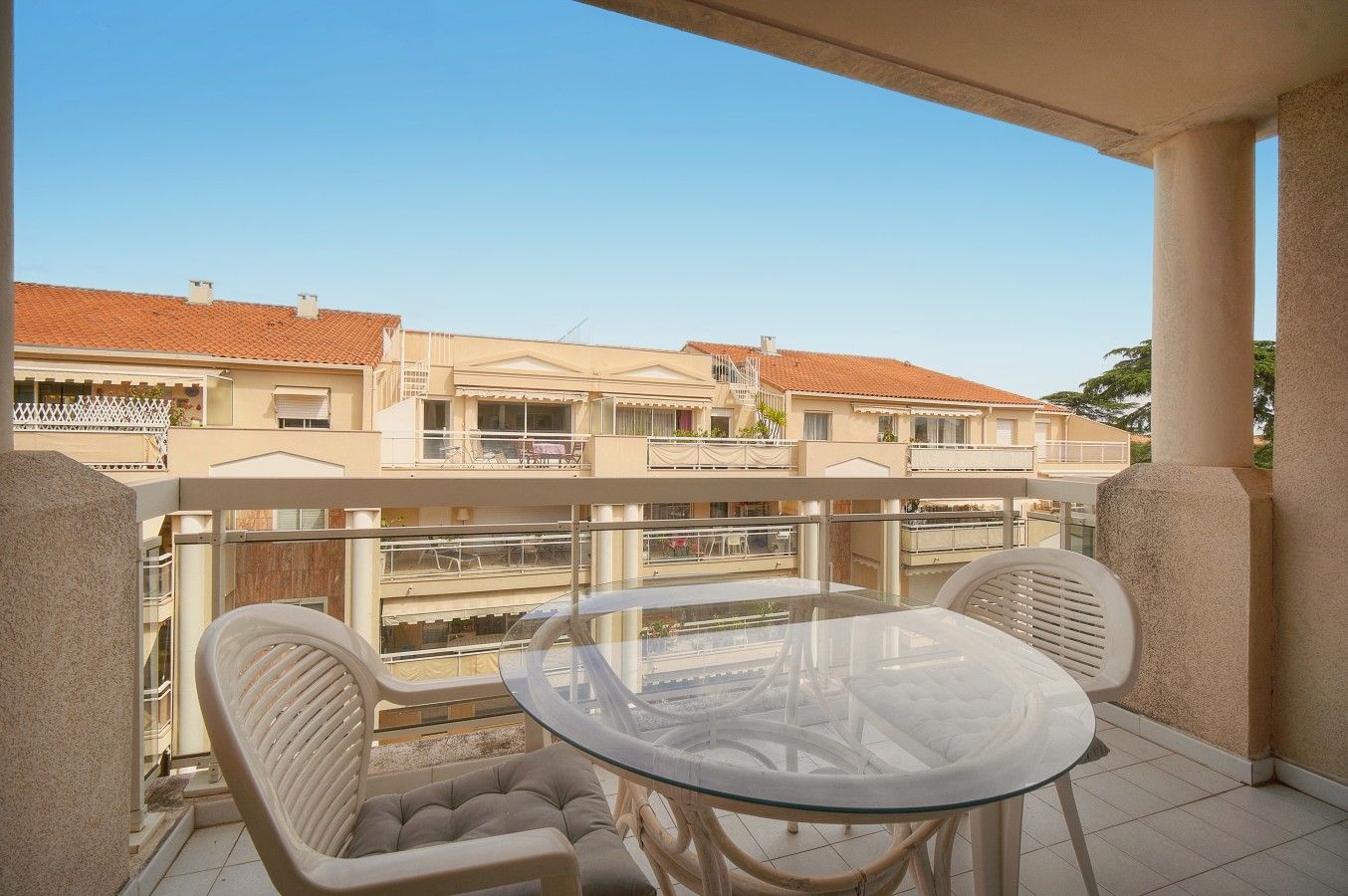 Un balcon avec une table en verre et deux chaises, donnant sur une rangée d’immeubles aux toits recouverts de tuiles rouges.