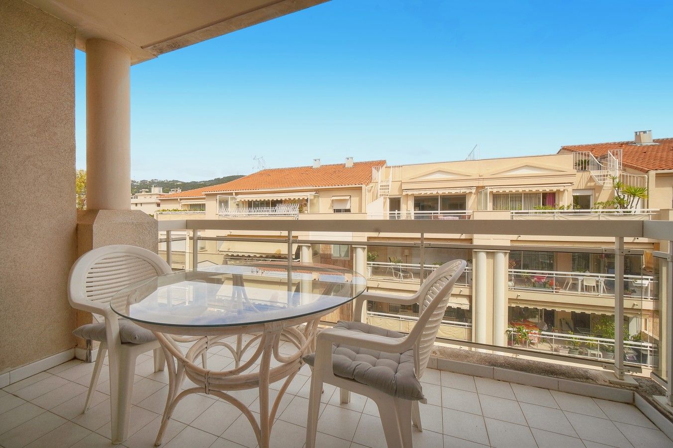 A balcony with a glass table and two chairs, overlooking a residential area with multiple buildings.