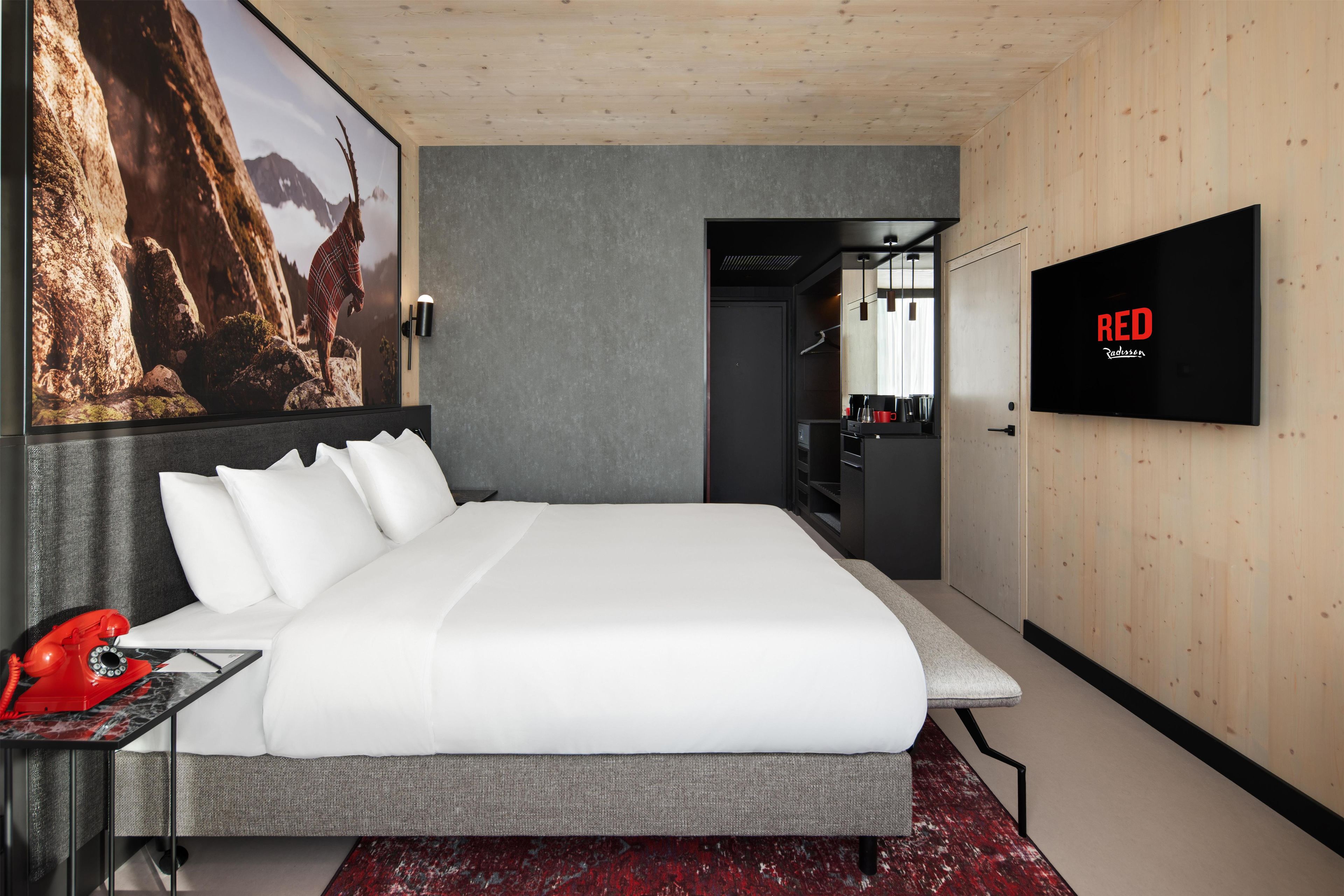 A modern hotel room with a large bed, a mounted TV, a red telephone, and a framed picture on the wall.