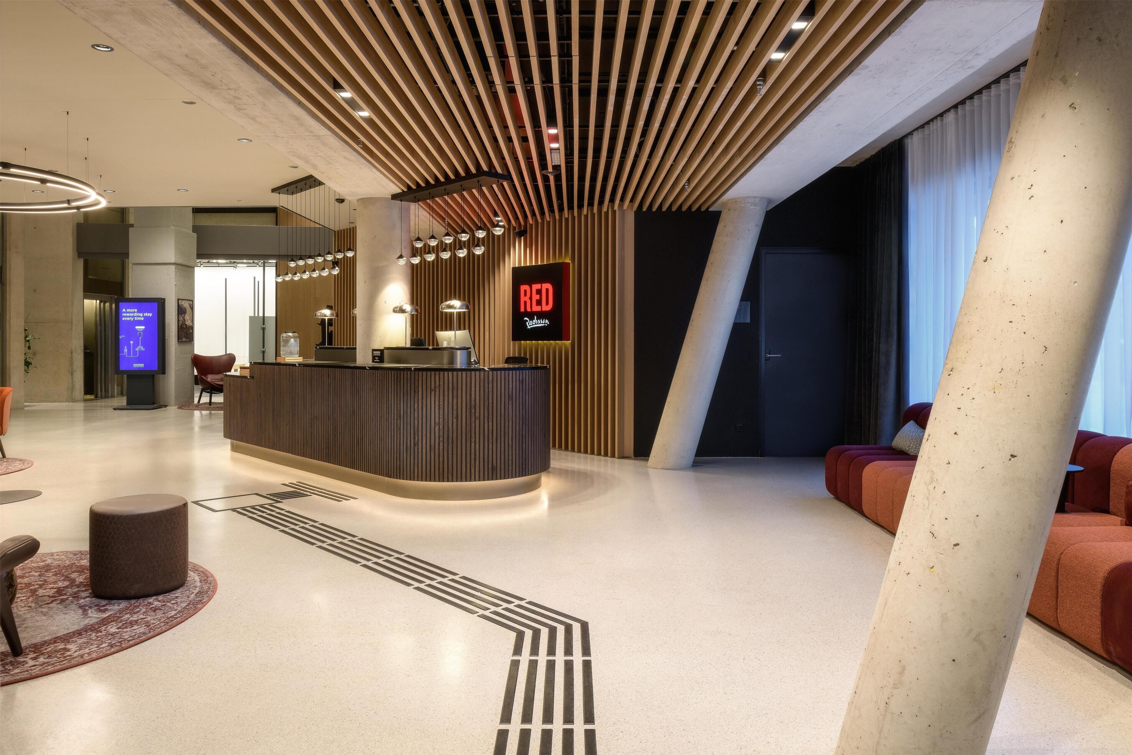 Eine moderne Hotelhalle mit Empfangstresen, Sitzgelegenheiten und einem großen Bildschirm, der 'RED' anzeigt.