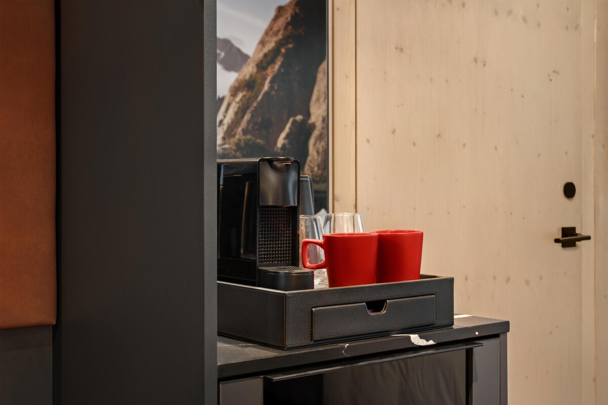 A coffee maker, two red cups, and a glass on a counter with a mountain view in the background.