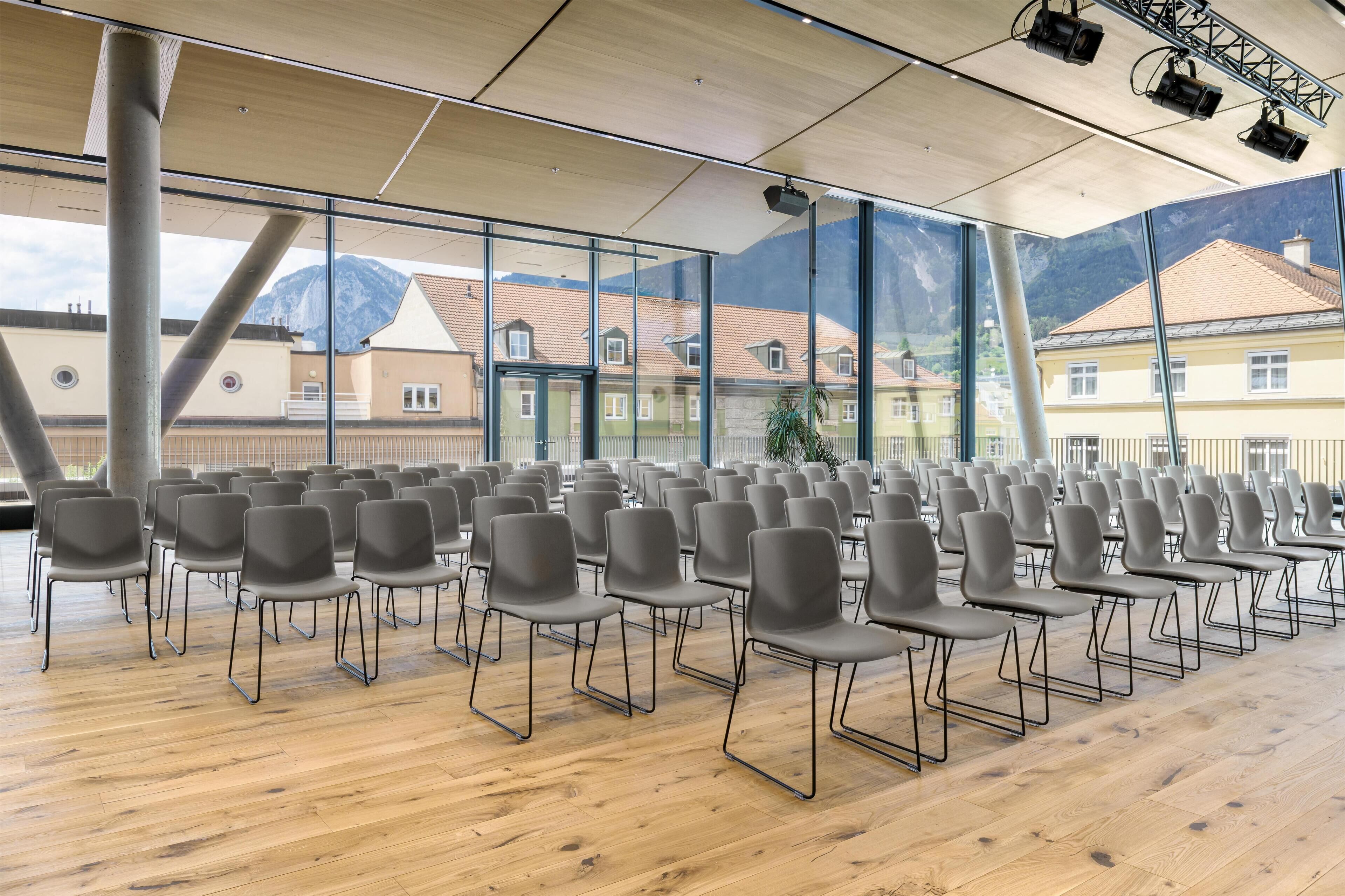 Un salón de conferencias con filas de sillas, ventanas amplias y suelo de madera.